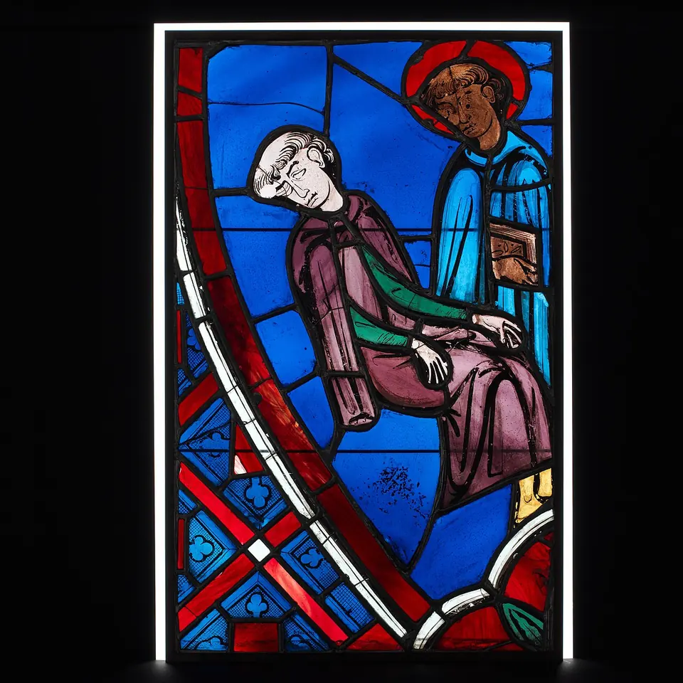 Medieval stained-glass panel in deep blue and red of a sleeping tonsured cleric leaning on a seat while a haloed figure holding a book stands beside him