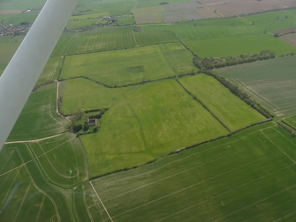 An aerial view of green fields with faint earthworks and a small church among trees, the outlines of the vanished priory visible in the grass