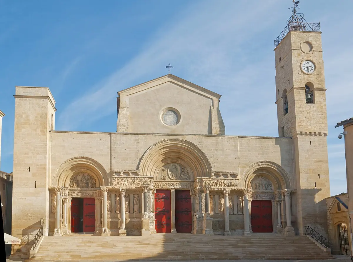 The pale stone Romanesque facade of the abbey church of Saint-Gilles with three arched portals and carved columns at the top of a wide flight of steps, beside a bell tower