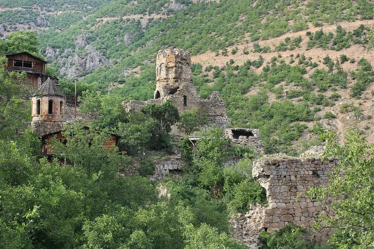 The ruined stone church and walls of Khandzta monastery among green forested mountains, with wooden village houses nearby