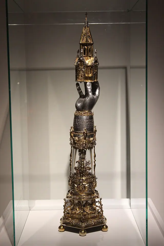A tall gilded silver reliquary shaped like an arm and hand rising from an ornate base and topped by a small Gothic tower, in a glass case