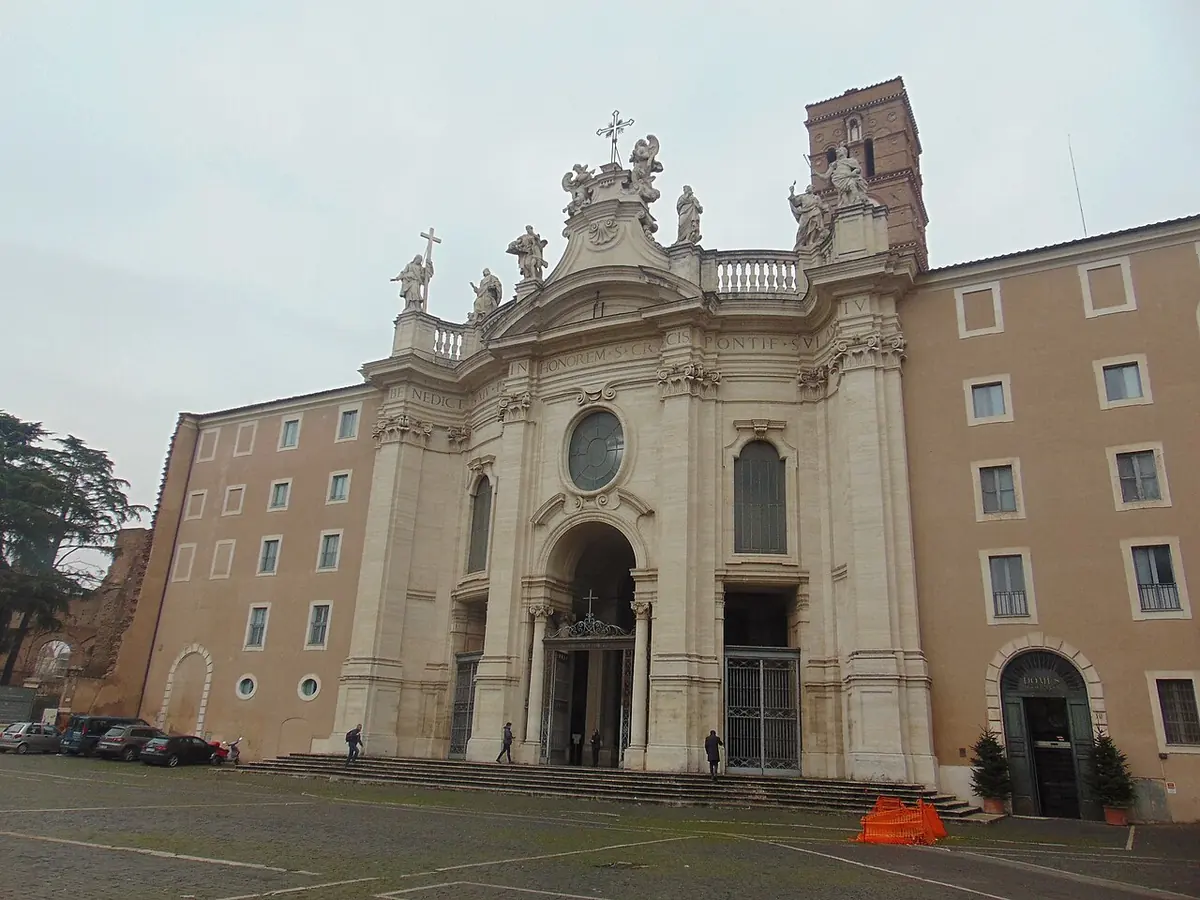 The white Baroque facade of Santa Croce in Gerusalemme with statues along its top and a Romanesque bell tower behind, beside a wide paved square