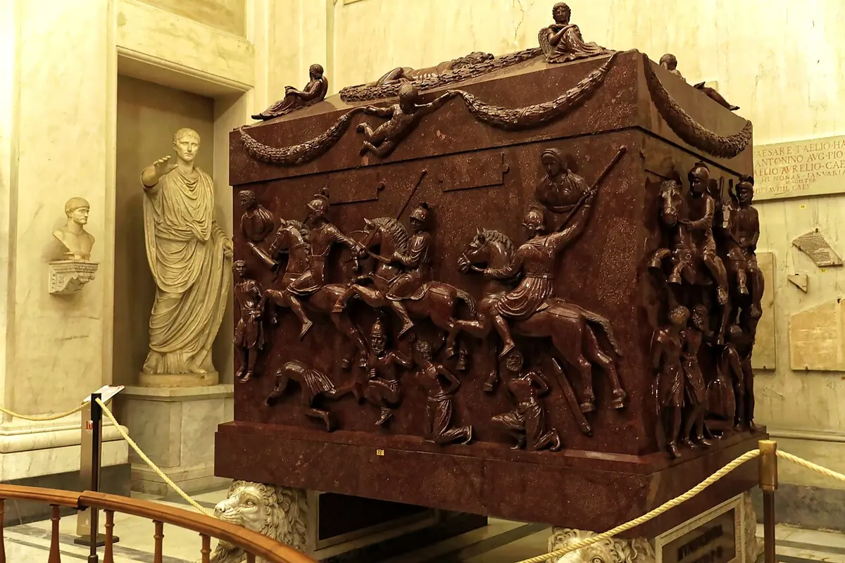 A huge red porphyry sarcophagus carved with Roman horsemen riding over fallen captives, with garlands and figures on its lid, in a marble museum hall