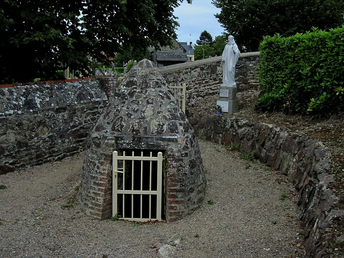 A small domed stone well house with an iron grille, set in a sunken garden path, with a white statue of St. Helier behind it