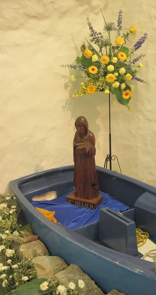 A carved wooden figure of a hooded hermit standing in a small blue boat, with summer flowers on a stand beside it, against a whitewashed wall