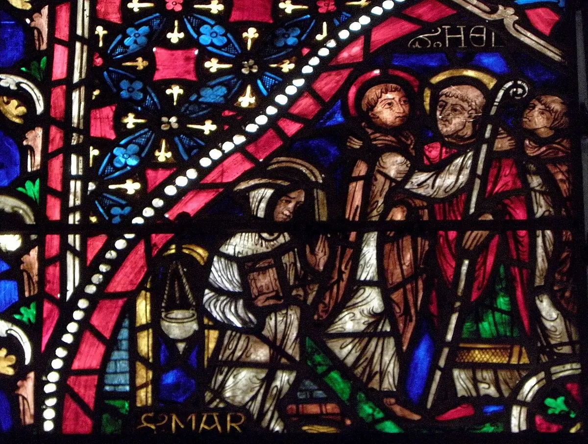 Medieval stained glass in deep reds and blues showing haloed monks and an abbot with a crozier greeting one another, with Latin lettering along the edges