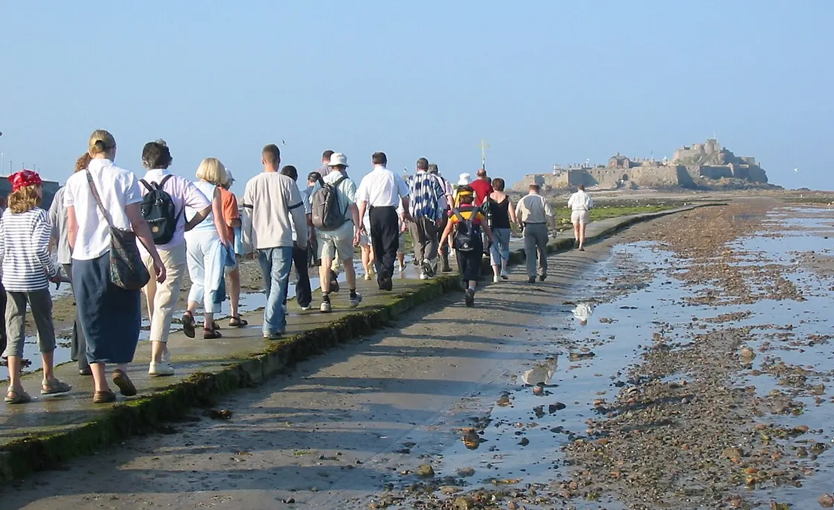 A long line of pilgrims walking across a wet causeway at low tide toward Elizabeth Castle under a clear sky