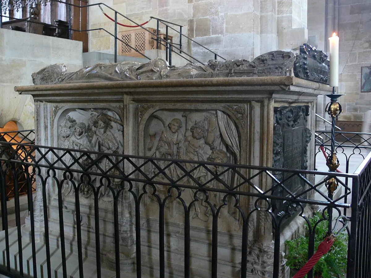 A large carved limestone tomb chest with relief panels and the figures of a crowned couple lying on top, behind an iron railing in a cathedral