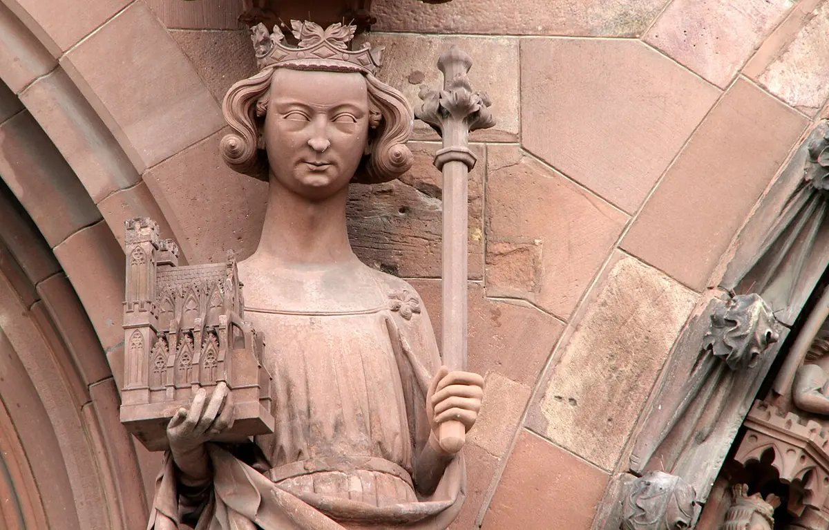 A red sandstone statue of a young crowned king holding a model of a church in one hand and a scepter in the other, set in a Gothic portal