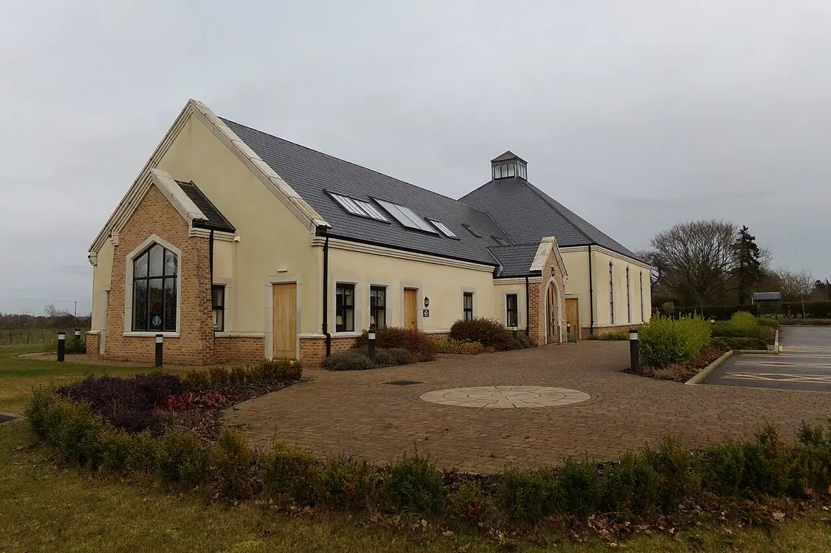 Modern cream-colored church with a slate roof, a small lantern on top and a tall arched window, behind a gravel forecourt and low hedges