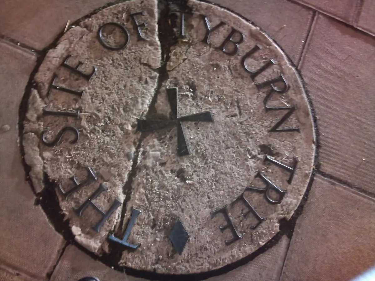 Round stone plaque set in a pavement with a small cross in the center and the words The Site of Tyburn Tree around the edge