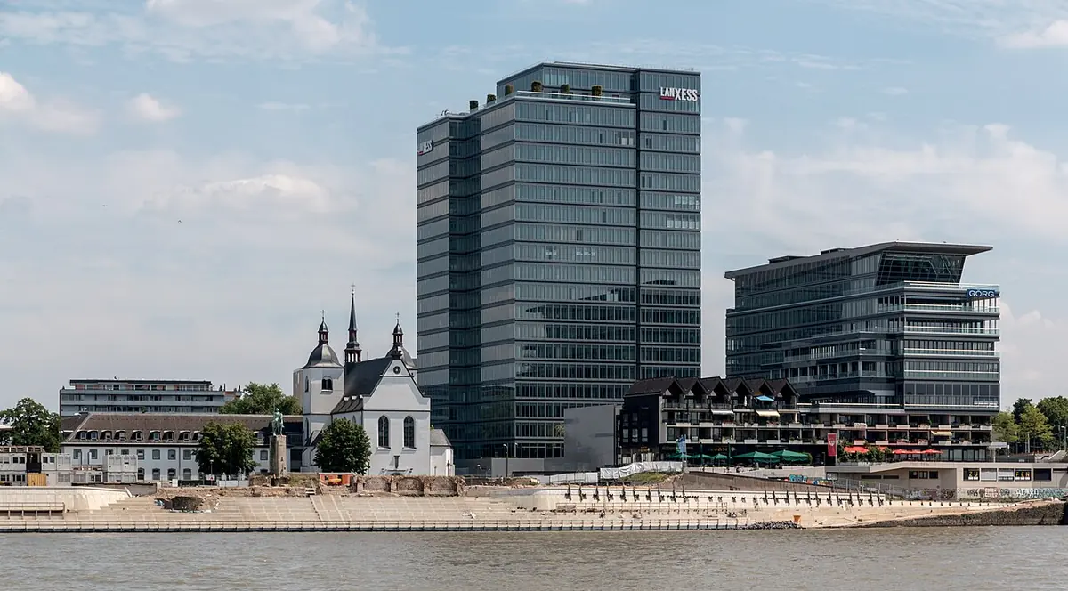 A white Baroque church with two small towers on the bank of the Rhine, with modern glass office towers rising behind it