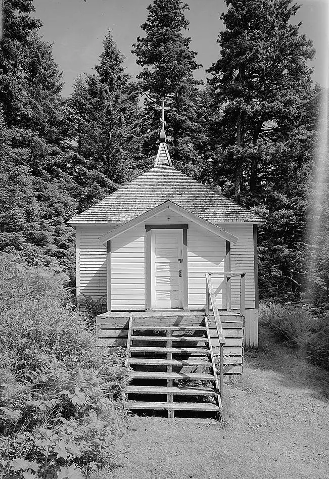 Black-and-white photograph of a small white wooden chapel with a little cupola and cross, steps leading up to its door, among tall spruce trees