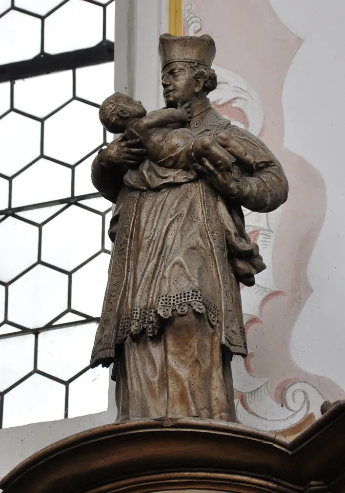 A carved wooden statue of a canon in a biretta and surplice holding the Christ Child in his arms, standing on a choir stall