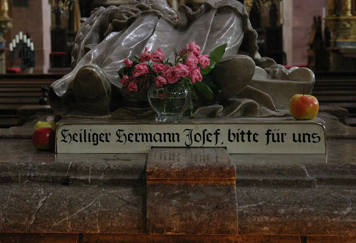 A marble tomb with a reclining figure, pink flowers in a glass vase and red apples placed on it, above a sign reading Heiliger Hermann Josef, bitte für uns