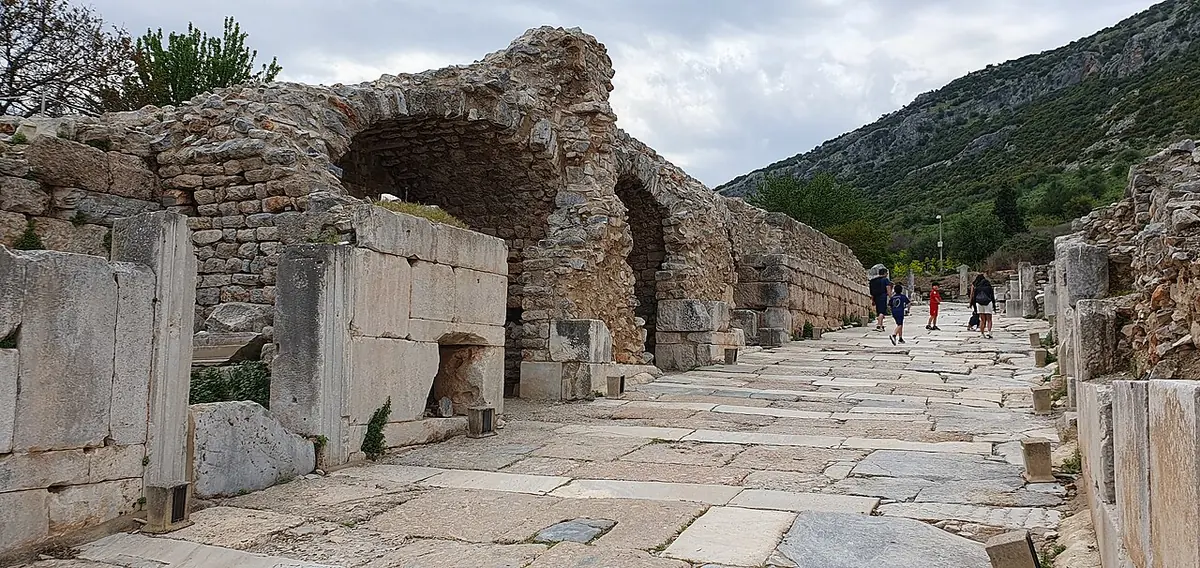 Marble-paved ancient street at Ephesus between ruined stone walls and arches, with visitors in the distance