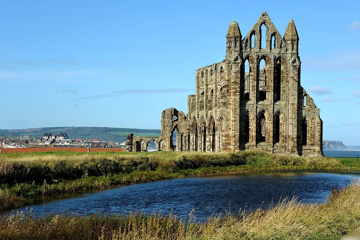 The tall Gothic ruins of Whitby Abbey on a grassy headland, reflected in a pond, with the North Sea and the town beyond