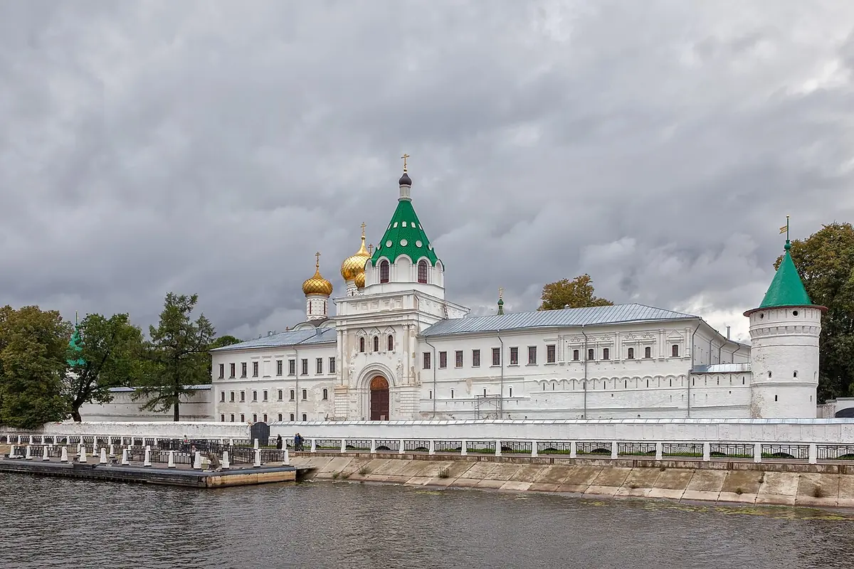 A long white monastery wall with towers and green and gold domes beside a river under a cloudy sky