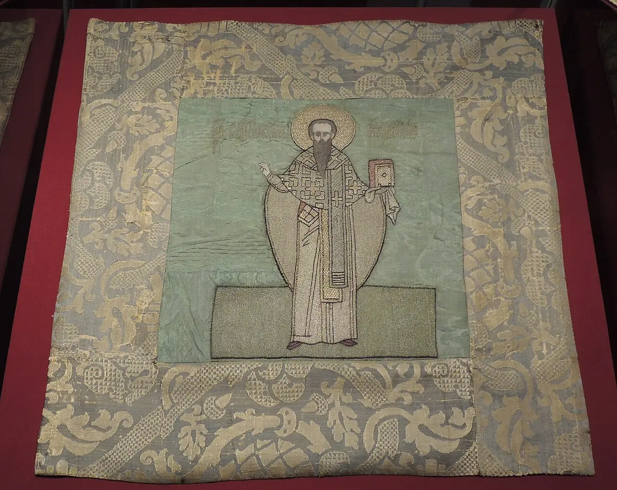A square silk cloth with a pale damask border and, in the center, an embroidered standing bishop holding a book