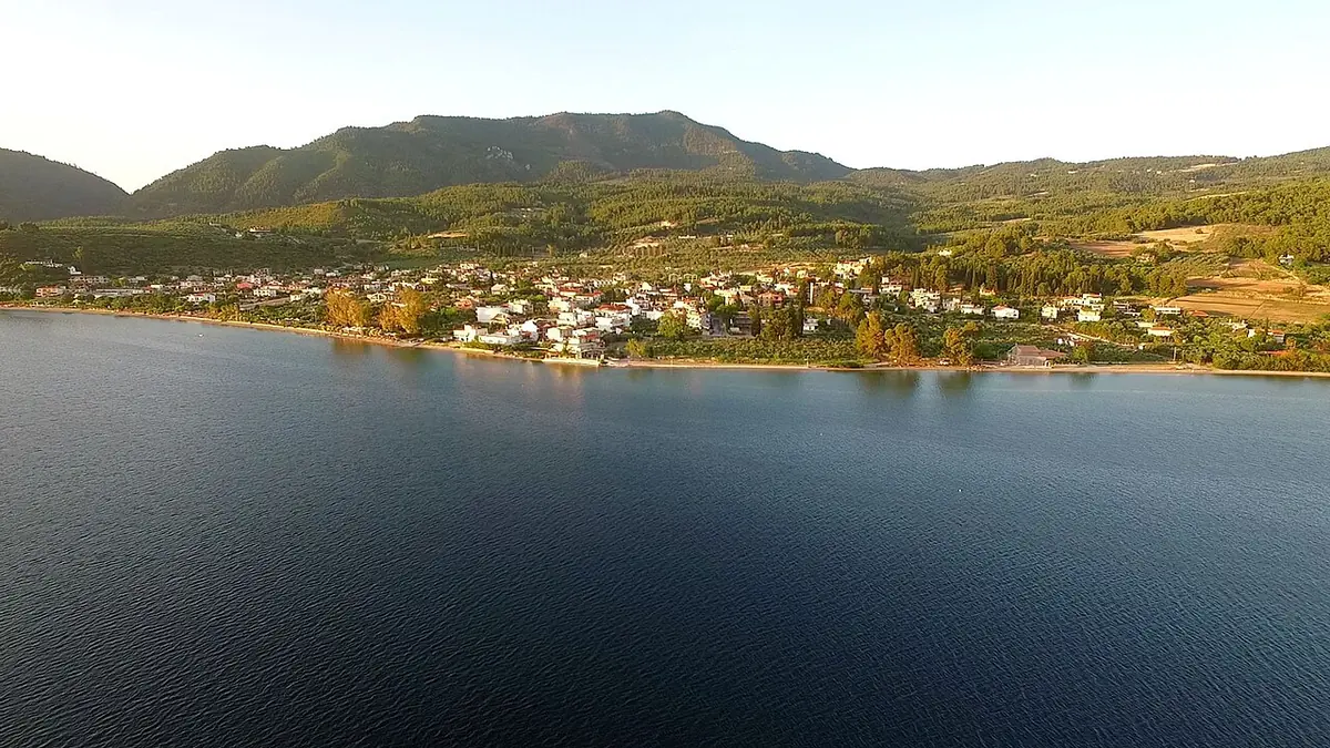 A small coastal village spread along a calm bay at sunset, with green wooded mountains rising behind it