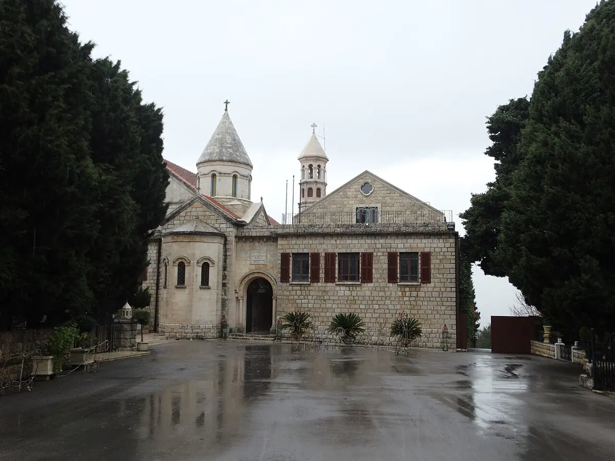 Stone monastery church with a conical-roofed dome and a bell tower, behind a wet courtyard flanked by dark cypress trees