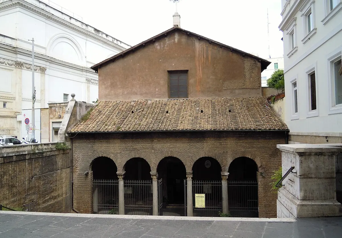 The plain brick facade of an early Christian church with a tiled roof and an arcaded porch of five arches, set below the level of the street