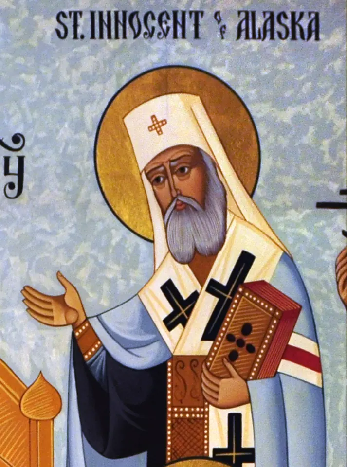 Modern icon of a white-bearded bishop with a gold halo, in a white klobuk and a pale blue omophorion marked with black crosses, holding a Gospel book and raising his hand, under the words St. Innocent of Alaska