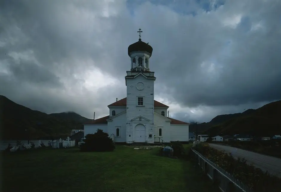 A white wooden church with a bell tower and green onion domes on a grassy flat, under dark clouds with mountains behind