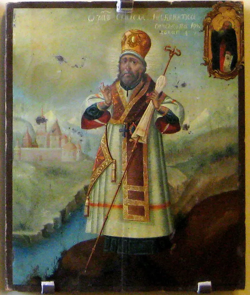 Painted icon of a bishop in a gold miter and white and gold vestments standing on a riverbank before a walled monastery, holding a staff and blessing