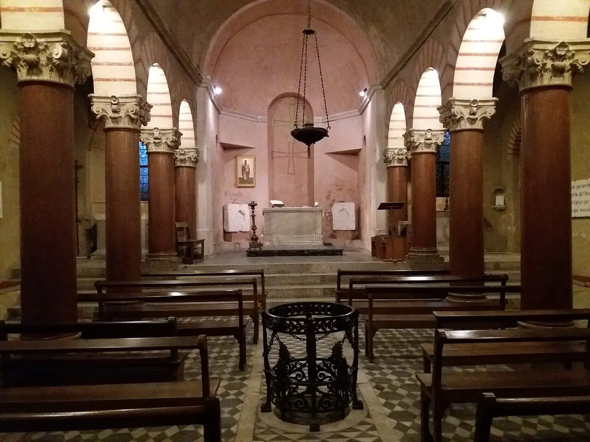 A crypt with red-brown columns and striped arches, wooden pews and a small altar in a pink apse, lit by a hanging lamp