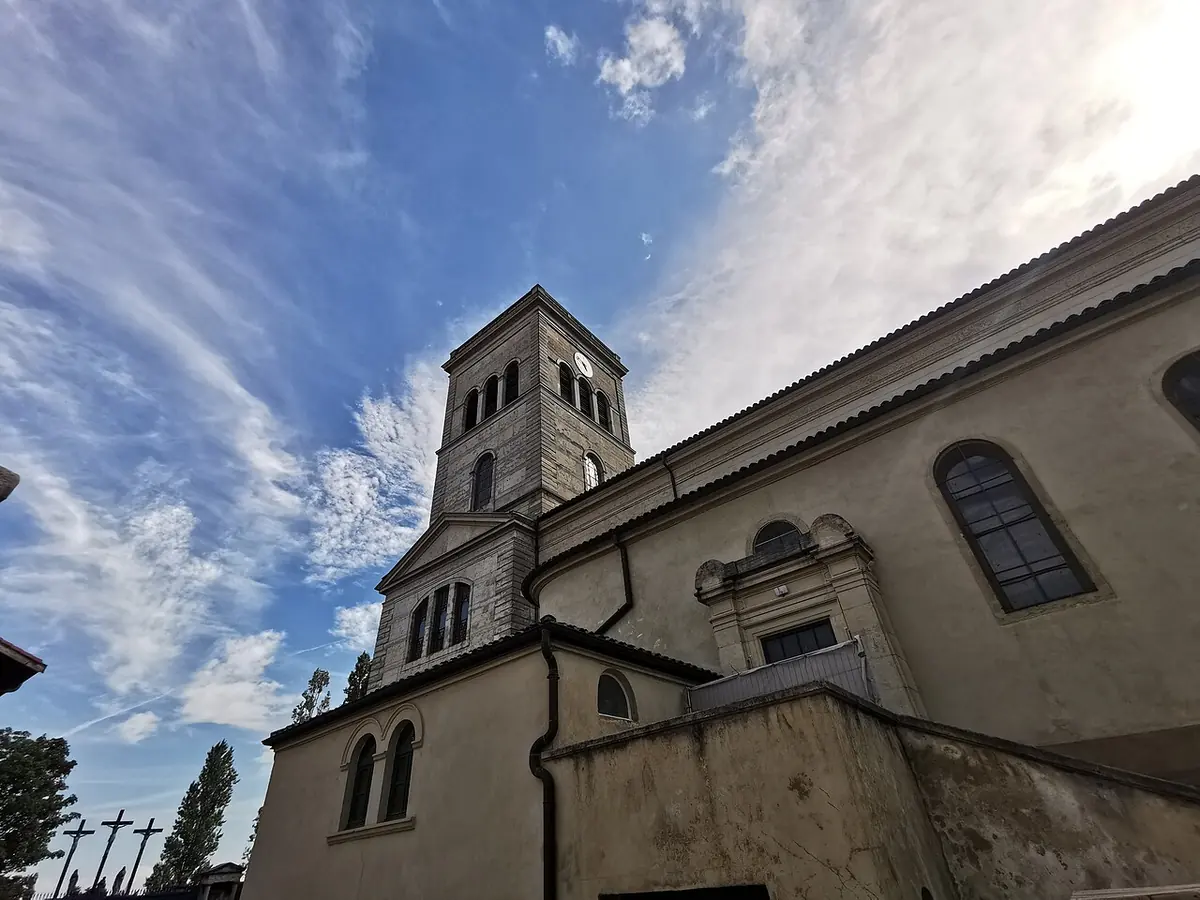 A pale stone church with a square bell tower seen from below against a cloudy blue sky, with the crosses of a calvary at the left