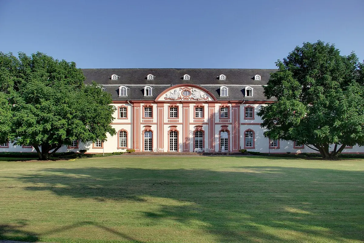 Long pink-and-white Baroque building with tall windows and a central pediment behind a green lawn and large trees under a blue sky