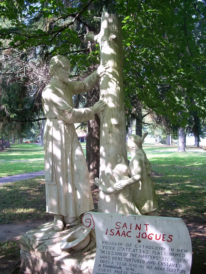 White statue of a Jesuit priest pointing to a cross on a tree trunk while two children kneel beside him, with a plaque reading Saint Isaac Jogues, under shady trees