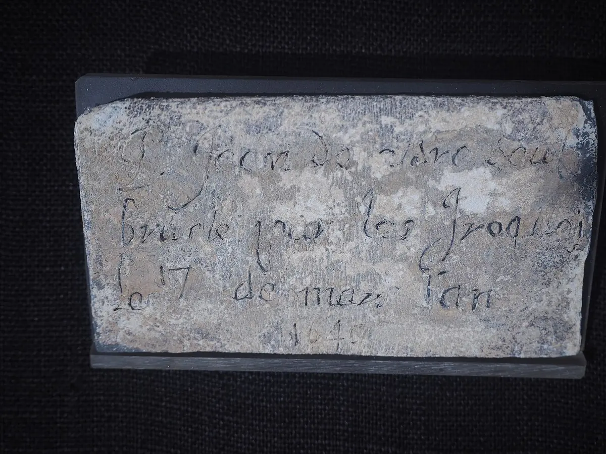 Small gray lead plate with a handwritten French inscription scratched into it, on a black background