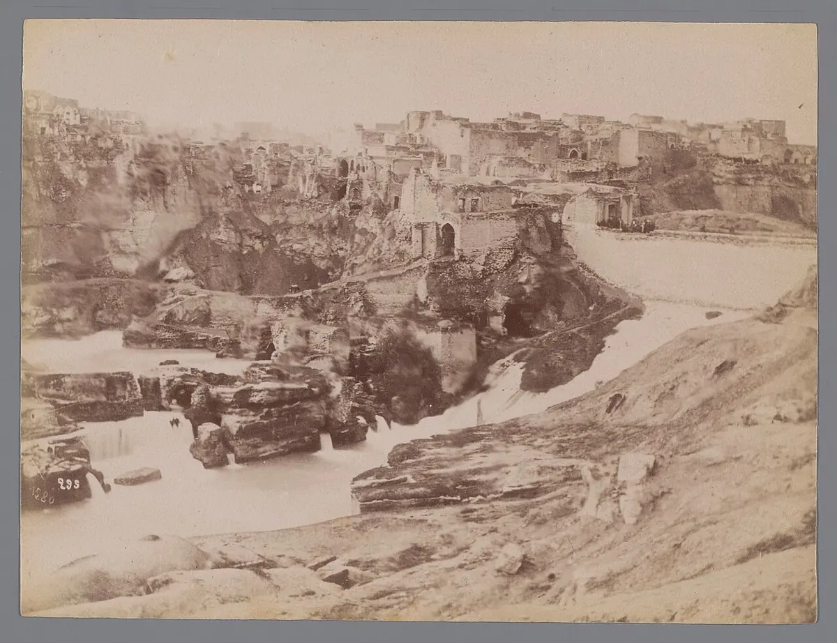 An old sepia photograph of a town of flat-roofed houses on a rocky bluff above a river with weirs and rough stone banks