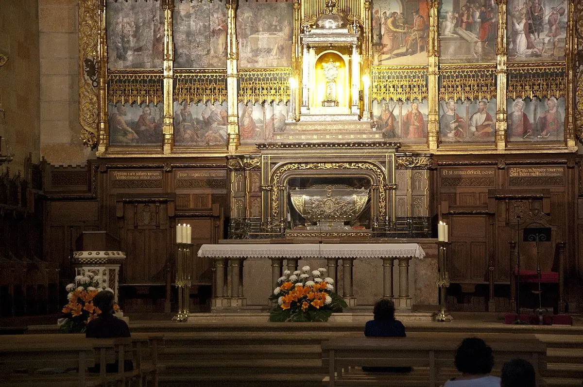 A church sanctuary with a large painted and gilded altarpiece, a silver reliquary chest above the altar and a few people praying in the pews