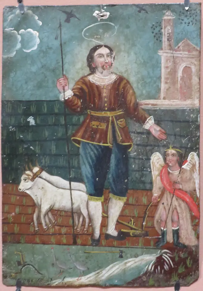 Mexican folk painting on tin of St. Isidore with a halo and a goad, standing in a field beside white oxen, with an angel holding the plow behind him and a church in the distance