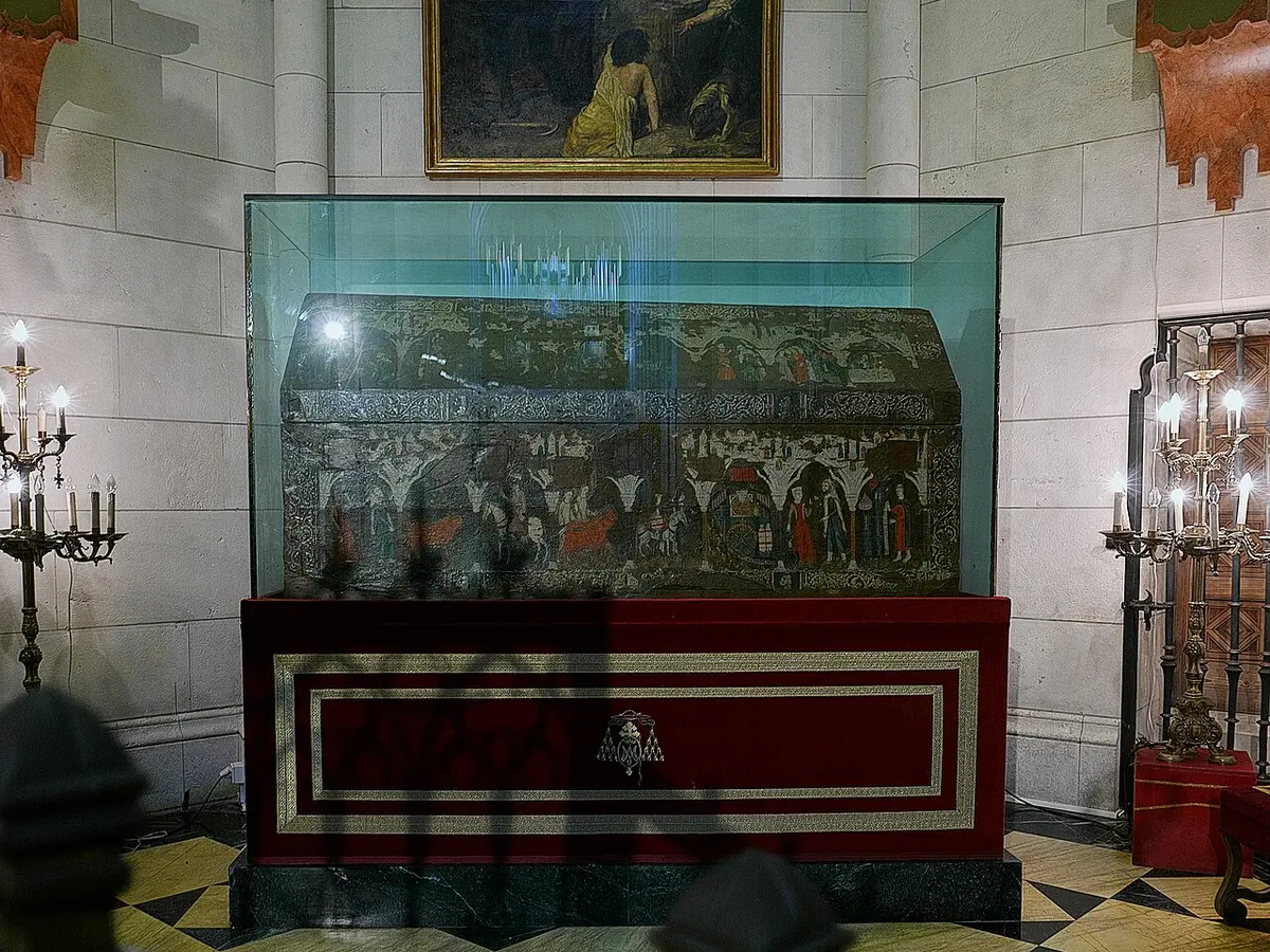 A painted wooden chest with small medieval scenes on its sides, displayed in a glass case on a red base in a chapel