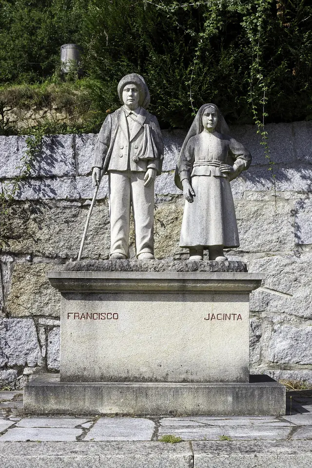 White stone statue of a boy in a cap holding a shepherd's crook beside a girl in a long dress and veil, on a pedestal carved with the names Francisco and Jacinta