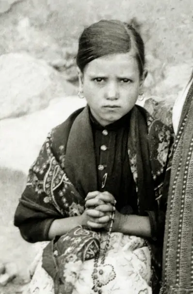Black-and-white photograph of a little girl in a patterned shawl with her hair parted in the middle, holding a rosary in her joined hands and looking straight at the camera