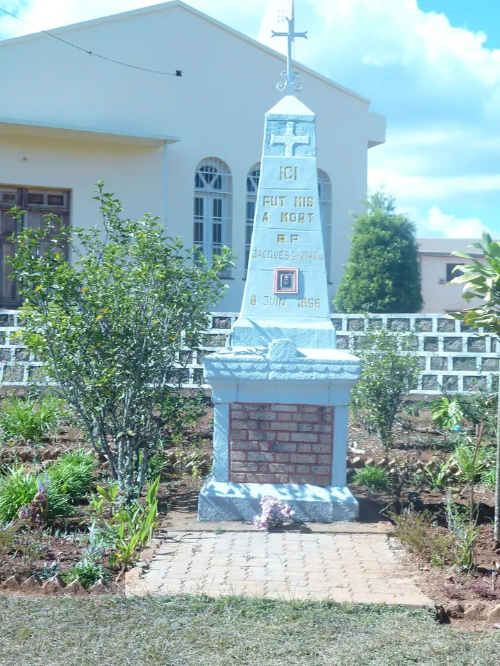 White obelisk topped by a cross with a French inscription and a small framed picture, standing in a garden in front of a white building