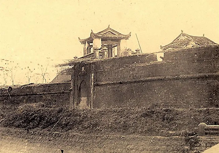Old sepia photograph of the thick brick walls of the Son Tay citadel with a gate tower topped by a curved tiled roof, overgrown with grass and bushes