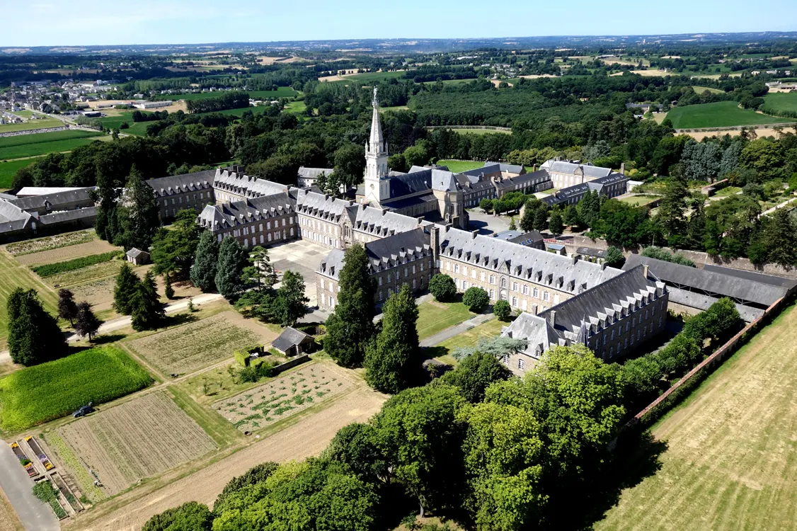 Aerial view of a large convent complex with a tall church spire, long wings of buildings and gardens among fields and woods