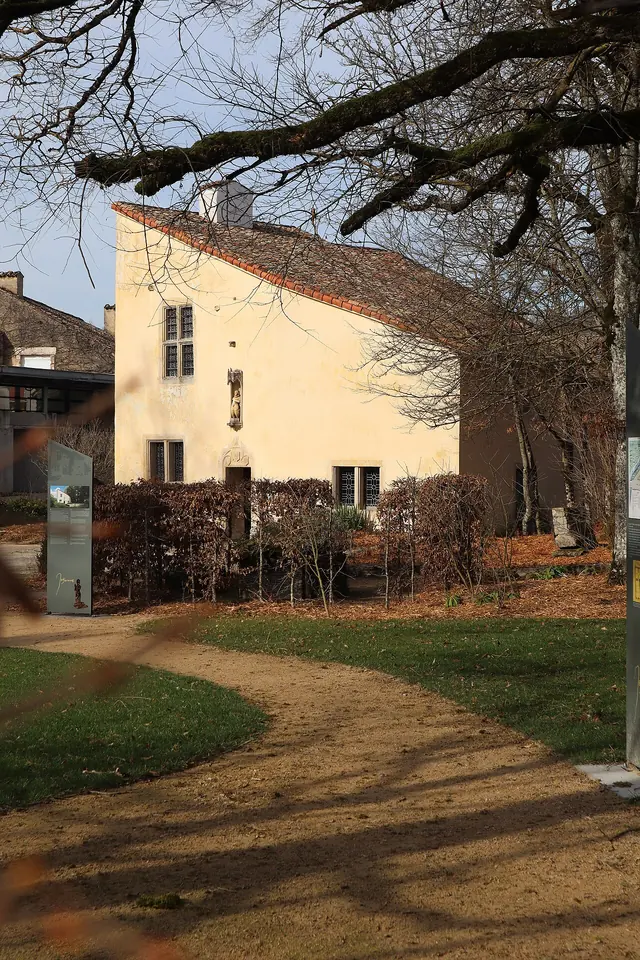 A small cream-colored stone house with a steep roof seen through bare trees at the end of a garden path