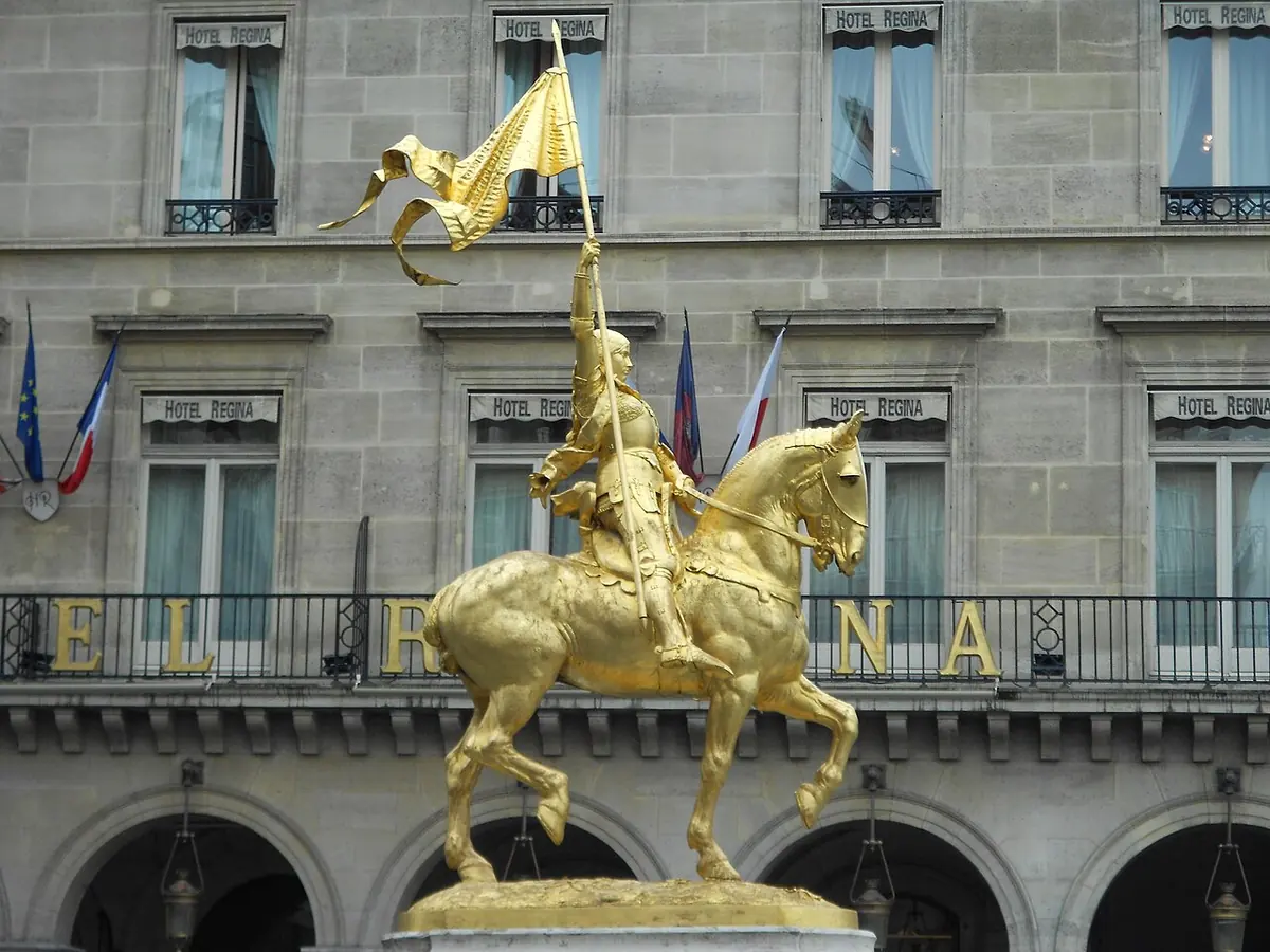 Gilded bronze statue of Joan in armor on horseback, holding a banner high, in front of a stone building with balconies
