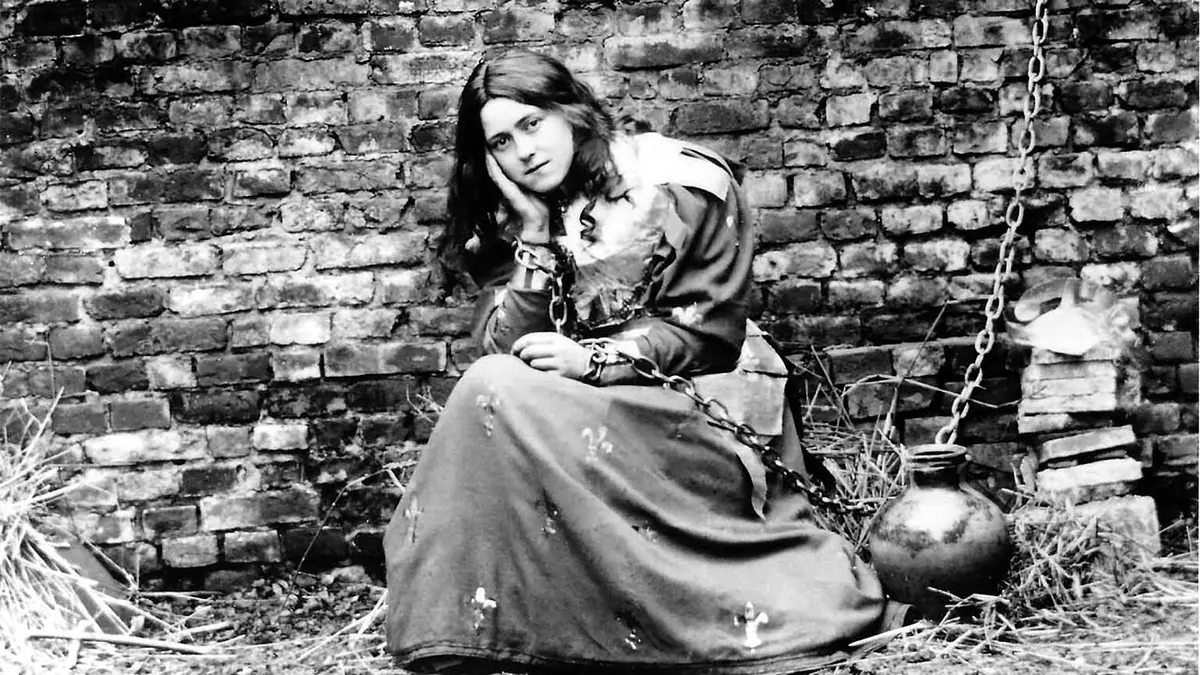 Black-and-white photograph of a young woman with long dark hair in a dress, seated on straw against a brick wall with a chain beside her, her head resting on her hand