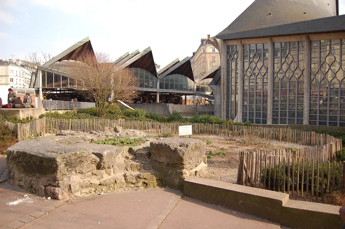Low stone foundations of a vanished church in a fenced garden on a town square, with the pointed slate roofs of a modern church and half-timbered houses behind