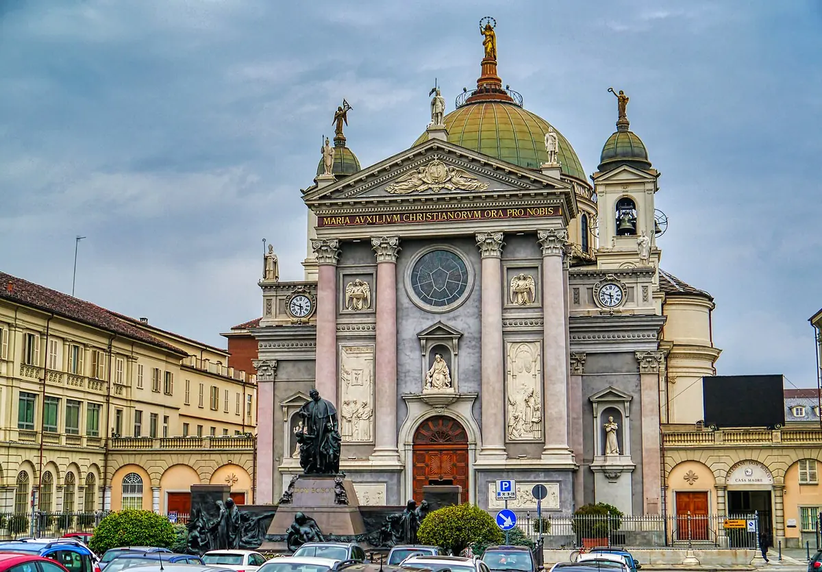 Pale stone church facade with columns, a rose window and a green dome topped by a gilded statue, with a monument in the square before it