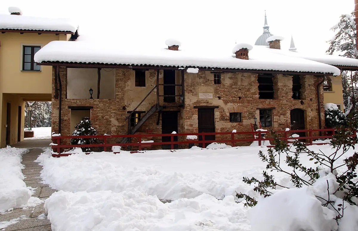 Small two-story brick farmhouse with a wooden balcony, its roof and yard covered with snow