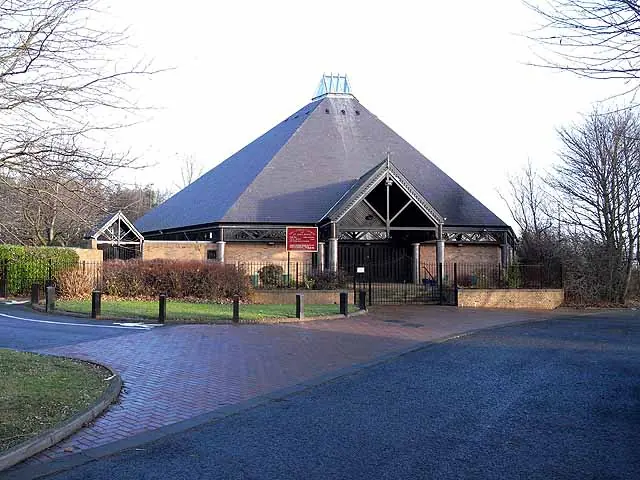 Modern church with a steep pyramid-shaped slate roof and brick walls, set among trees beside a curving road